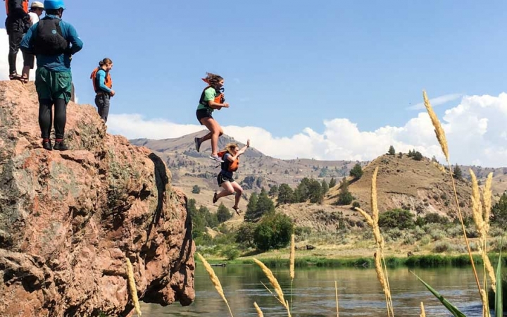 two outward bound students wearing life jackets jump off of a rocky overlook into water while others look on